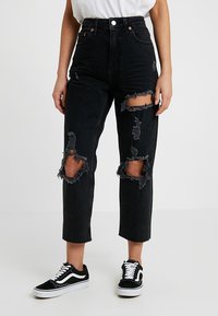 Jeans neri a vita alta con tessuto strappato e grandi dettagli strappati su entrambe le ginocchia. Presentano una gamba dritta e orlo sfilacciato. Abbinati a sneakers nere.