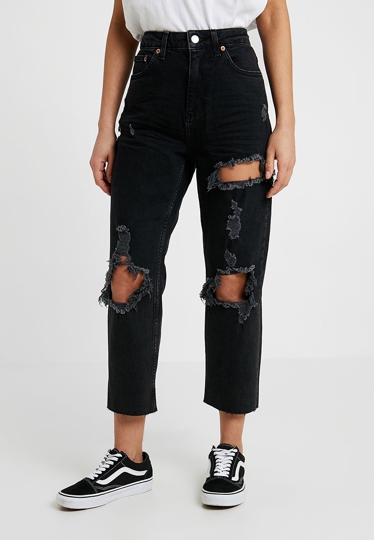 Jeans neri a vita alta con tessuto strappato e grandi dettagli strappati su entrambe le ginocchia. Presentano una gamba dritta e orlo sfilacciato. Abbinati a sneakers nere.