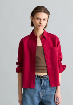 Junge Person mit kurzen braunen Haaren trägt ein offenes rotes Hemd über einem braunen Tanktop und blauen Jeans mit einem schwarzen Gürtel.