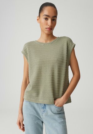 Donna con capelli raccolti che indossa una maglietta verde oliva a maniche corte con texture e jeans azzurri, in piedi davanti a uno sfondo semplice.