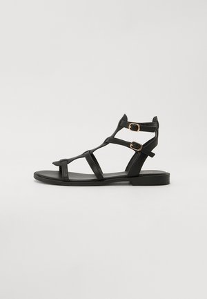 Sandales strappy noires en cuir avec semelle plate, deux boucles sur les brides de cheville, design ouverte à l'avant et silhouette minimaliste.