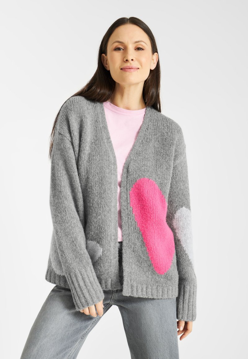 Grijze gebreide cardigan met roze en witte hartpatronen, een open voorkant, geribbelde manchetten en twee zijzakken. Draagt over een roze shirt.