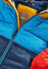 Giacca isolante colorata con pannelli blu, blu navy e rossi, chiusura frontale con zip, gocce d'acqua sul tessuto e una fodera interna gialla.