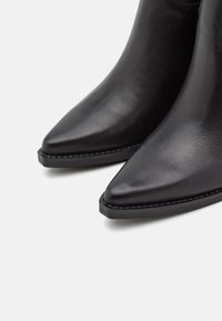 Botas de cuero negro hasta el tobillo con punteras puntiagudas y una textura suave. Presenta una costura contrastante a lo largo de la suela para un detalle adicional.