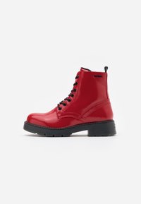 Botas de tobillo de charol rojas con cordones negros, punta redonda y suela gruesa negra. Incluyen una tira en el talón para facilitar su uso.