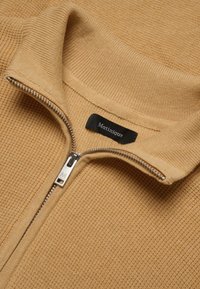Pull beige zippé en tissu texturé, col côtelé, fermeture éclair argentée et étiquette noire affichant le nom de la marque, "Matinique".