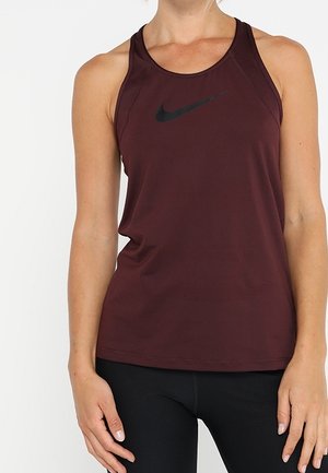 Donna che indossa una canotta sportiva bordeaux senza maniche Nike e leggings neri, in piedi con le braccia rilassate lungo i fianchi.
