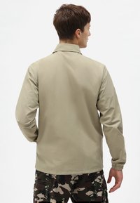 Lys beige jakke med krage, lange ermer og elastiske mansjetter, med en glatt tekstur. Brukt over kamuflerte cargo-bukser.