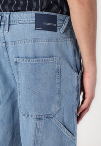 Pantalons en denim bleu clair avec une poche à rabat ornée d'une couture en zigzag et une étiquette de marque marine sur la taille.