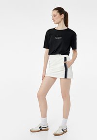 Zwarte T-shirt met korte mouwen en witte tekst, gecombineerd met een witte minirok met een marineblauwe streep, en witte sneakers met zwarte accenten.