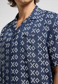Chemise à manches courtes en tissu bleu foncé avec un motif géométrique blanc, dotée d'une poche poitrine et d'une étiquette de marque sur la poche.