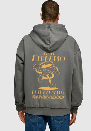 Graue Kapuzenjacke mit einem großen gelben Grafikdruck auf dem Rücken, der eine Cartoonfigur mit einer Tasse und dem Text "MEHR ESPRESSO WENIGER DEPRESSE" zeigt.