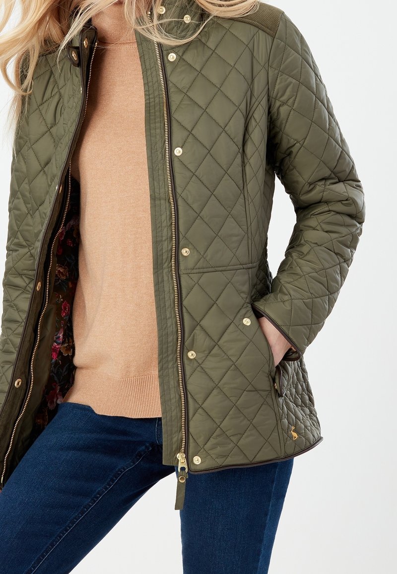 Gequilted olijfgroene jas met een ritssluiting, gouden hardware en een bloemenvoering; gecombineerd met een beige trui en blauwe spijkerbroek.