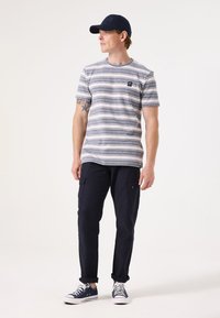 Gestreepte t-shirt met korte mouwen in marineblauw en wit, gecombineerd met donkere marineblauwe cargo pants. Een blauwe pet en witte sneakers maken de outfit af.