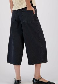 Pantalon en denim noir à jambes larges avec poches arrière, ourlet effiloché et coupe décontractée, associé à des ballerines noires et un haut en tricot léger.