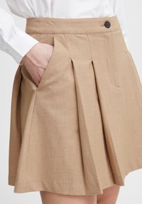 Plisseret skjørt i lys beige stoff med en glatt tekstur. Har en enkelt knappelukking og sidelommer for funksjonalitet.