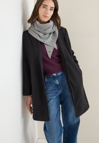 Cappotto lungo nero sopra un maglione viola scuro, abbinato a jeans azzurri chiari. Una sciarpa grigia a fantasia avvolge il collo. Sfondo neutro.