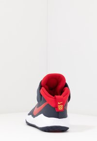 Nike-kengät, joissa on tummansininen päällinen, punainen verkkomallinen kaulus ja valkoinen välipohja. Huomionarvoinen punainen logo ja tekstuuripinnat kantapäässä.