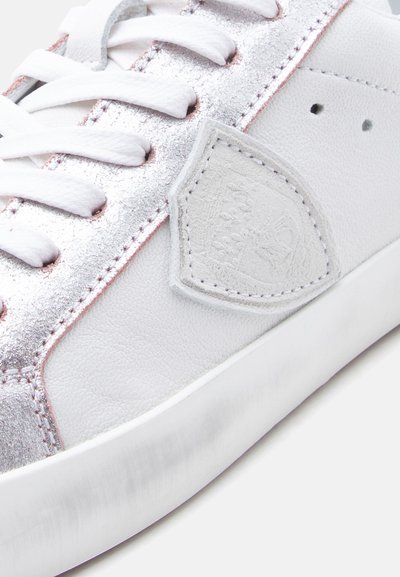 Λευκό αθλητικό παπούτσι με μεταλλικές ασημί λεπτομέρειες, υφή από δέρμα και ροζ ραφές. Διαθέτει λευκά κορδόνια και λεπτομέρεια με λογότυπο ασπίδας.