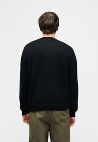 Homme de dos portant un pull à manches longues noir et un pantalon cargo vert olive sur un fond blanc uni.
