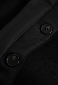 Gros plan sur un tissu noir avec deux boutons noirs à quatre trous cousus sur la zone du col d’un manteau ou d’une veste.