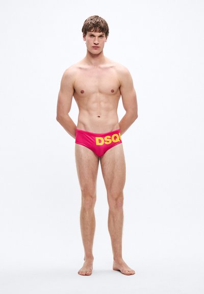 Rožinės plaukimo glaudės su dideliu geltonu „DSQ“ logotipu ant juosmens juostos. Lygus tekstūra ir prigludęs dizainas. Modelis stovi basomis.