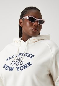 Fehér kapucnis pulóver, amelyen sötétkék hímzett szöveg olvasható: "HILFIGER est 1985 NEW YORK." Rózsaszín napszemüveg keskeny kerettel. Texturált anyag.