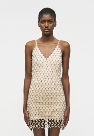 Frau trägt ein ärmelloses beiges Minikleid mit einer großen netzartigen Perlenüberlagerung, steht vor einem schlichten weißen Hintergrund.