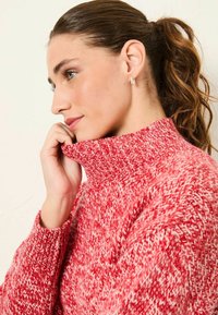 Roter gestrickter Pullover mit hohem, geripptem Rollkragen, der ein strukturiertes Muster aufweist. Das Model berührt den Kragen und schaut leicht zur Seite.