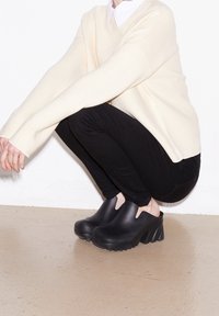 Schwarze Wedge-Pantoletten mit ausgeschnittenen Seiten, aus glattem Material. Mit einer dicken, geriffelten Sohle und einer abgerundeten Zehenpartie vor einem neutralen Hintergrund.