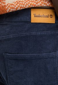 Pantalones de pana azul marino con un bolsillo trasero, que presentan una etiqueta de cuero color marrón de Timberland cosida en la cintura. Detalle de tejido texturizado visible.
