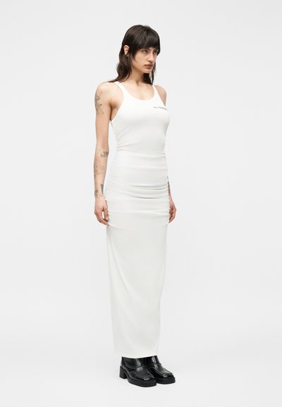 Γυναίκα με σκούρα μαλλιά και τατουάζ, φορώντας εφαρμοστό λευκό αμάνικο φόρεμα maxi και μαύρα παπούτσια με πλατφόρμα, στέκεται μπροστά σε απλό φόντο.