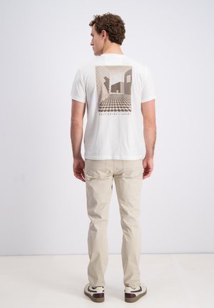 Man draagt een wit T-shirt met geometrisch gebouwontwerp en beige broek, staand voor een effen witte achtergrond.