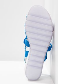 Sandale bleue avec une tige texturée et plusieurs sangles, dotée d'une semelle blanche à rainures et d'une semelle extérieure en caoutchouc transparent.