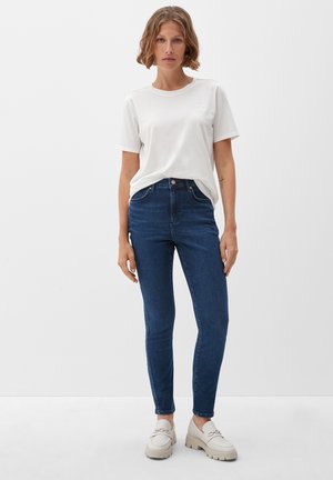 Vrouw in een witte korte mouwen t-shirt, donkere skinny jeans en beige chunky loafers, staand tegen een effen witte achtergrond.