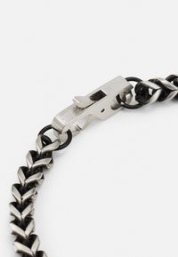 Bracelet en chaîne en métal argenté avec des sections entrelacées et un accent circulaire noir. Dispose d'un fermoir à finition polie avec marquage.