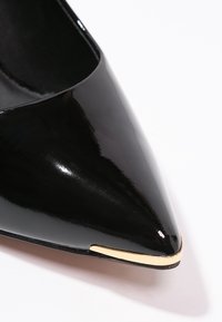 Chaussure pour femme noire brillante à bout pointu avec une fine bordure dorée métallique le long du bord avant, sur fond blanc.