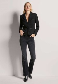 Schwarzer strukturierter Blazer mit V-Ausschnitt, kombiniert mit schwarzen Jeans, die am Saum ein allmähliches Nietenmuster aufweisen. Spitz zulaufende schwarze Stiefel.