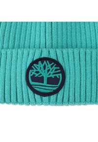 Bonnet côtelé turquoise avec un patch circulaire bleu marine arborant un logo d'arbre stylisé en broderie verte, texture douce.