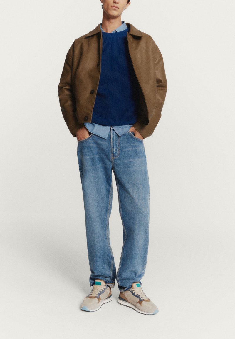 Giacca di pelle marrone, maglione blu a maglia, camicia azzurro chiaro, jeans azzurro chiaro larghi e scarpe beige con dettagli in teal.