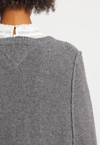 Grå stickad tröja med rund halsringning, strukturerat mönster, synliga sömmar och en avslappnad passform, visar baksidan och axelområdet.