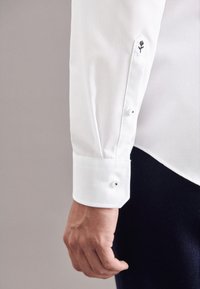 Camicia bianca a maniche lunghe con polsini abbottonati decorati da una rosa ricamata nera. Tessuto morbido, vestibilità strutturata e dettagli discreti sui polsini.