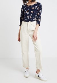 Marineblaue Bluse mit Blumenmuster und drei Viertelärmeln, kombiniert mit cremefarbenen, hochgeschnittenen Jeans und weißen Sneakers mit blauen und roten Akzenten.