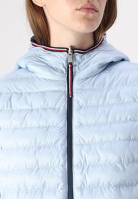 Lichtblauwe pufferjack met een capuchon, voorzien van horizontale gestikte naden, een marineblauwe rits en rode en witte accentstrepen op de kraag.