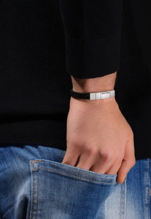 Bracciale in pelle nera con dettagli argentati, elementi metallici rettangolari, texture liscia, indossato su un polso, abbinato a jeans in denim.