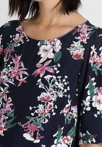 Blusa floreale blu navy con maniche corte, caratterizzata da fiori multicolori nei toni di rosa, bianco e verde, e scollatura rotonda.