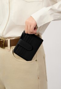 Portafoglio in pelle nera con finitura testurizzata, dotato di slot per carte e di un compartimento con zip, parzialmente visibile in una tasca di pantaloni beige.