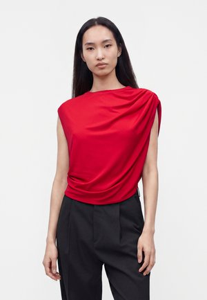 Jeune femme aux longs cheveux foncés portant un haut rouge sans manches drapé et un pantalon noir taille haute sur un fond clair uni.