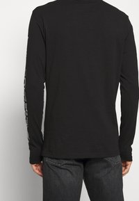 Svart långärmad tröja med rund halsringning, prydd med vit grafisk text längs den högra ärmen. Tillverkad av ett mjukt och lätt material.