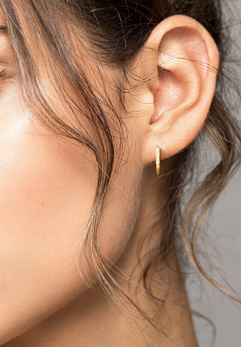 Goldene Creolen mit strukturiertem Oberflächen, kleines und dünnes Design, getragen am Ohr in Nahaufnahme. Haare bedecken teilweise das Ohrläppchen.